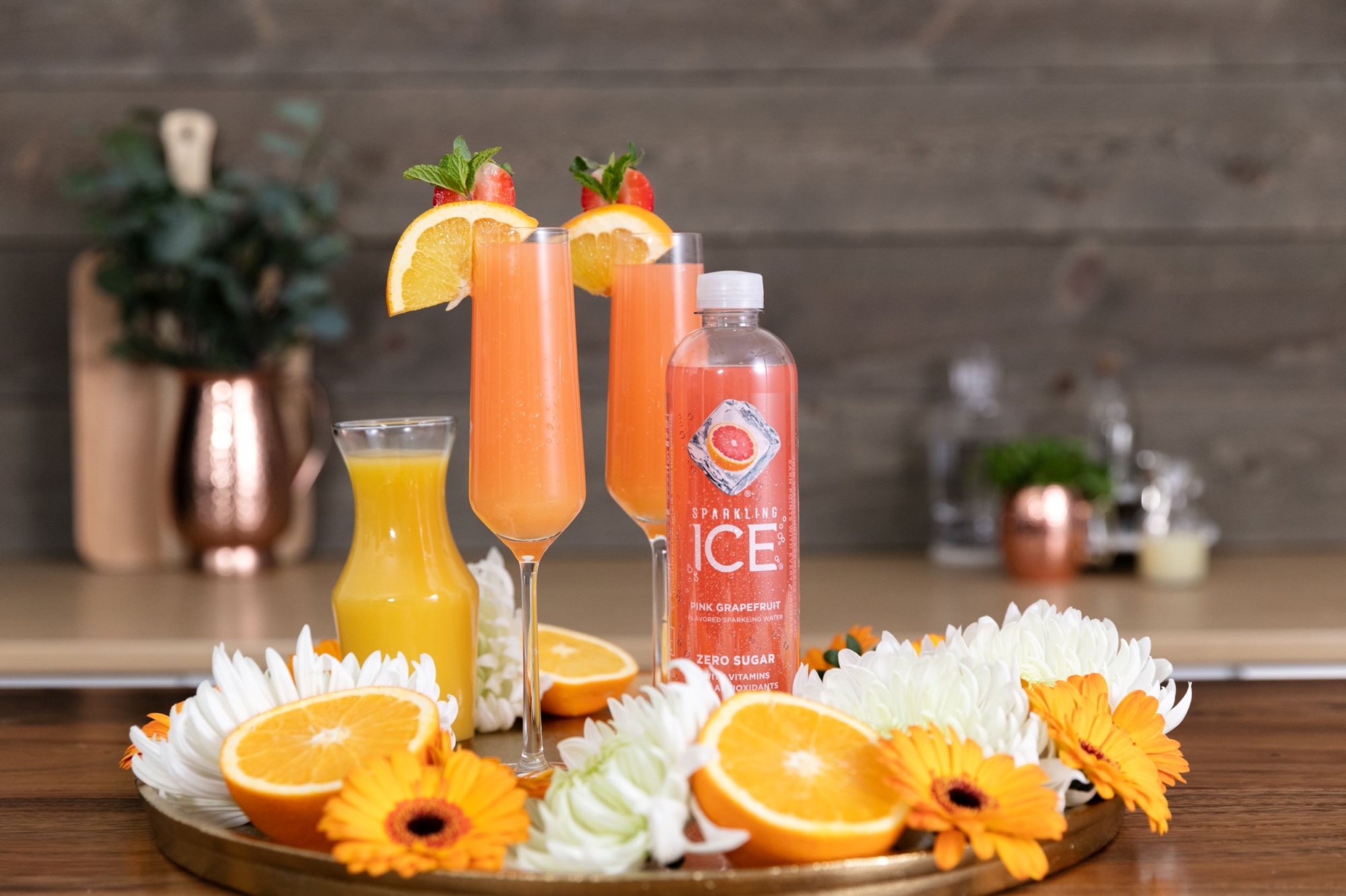 Sparkling Ice Pink Grapefruit mockmosa in a tall glass on table surrounded by fresh fruit and flowers.